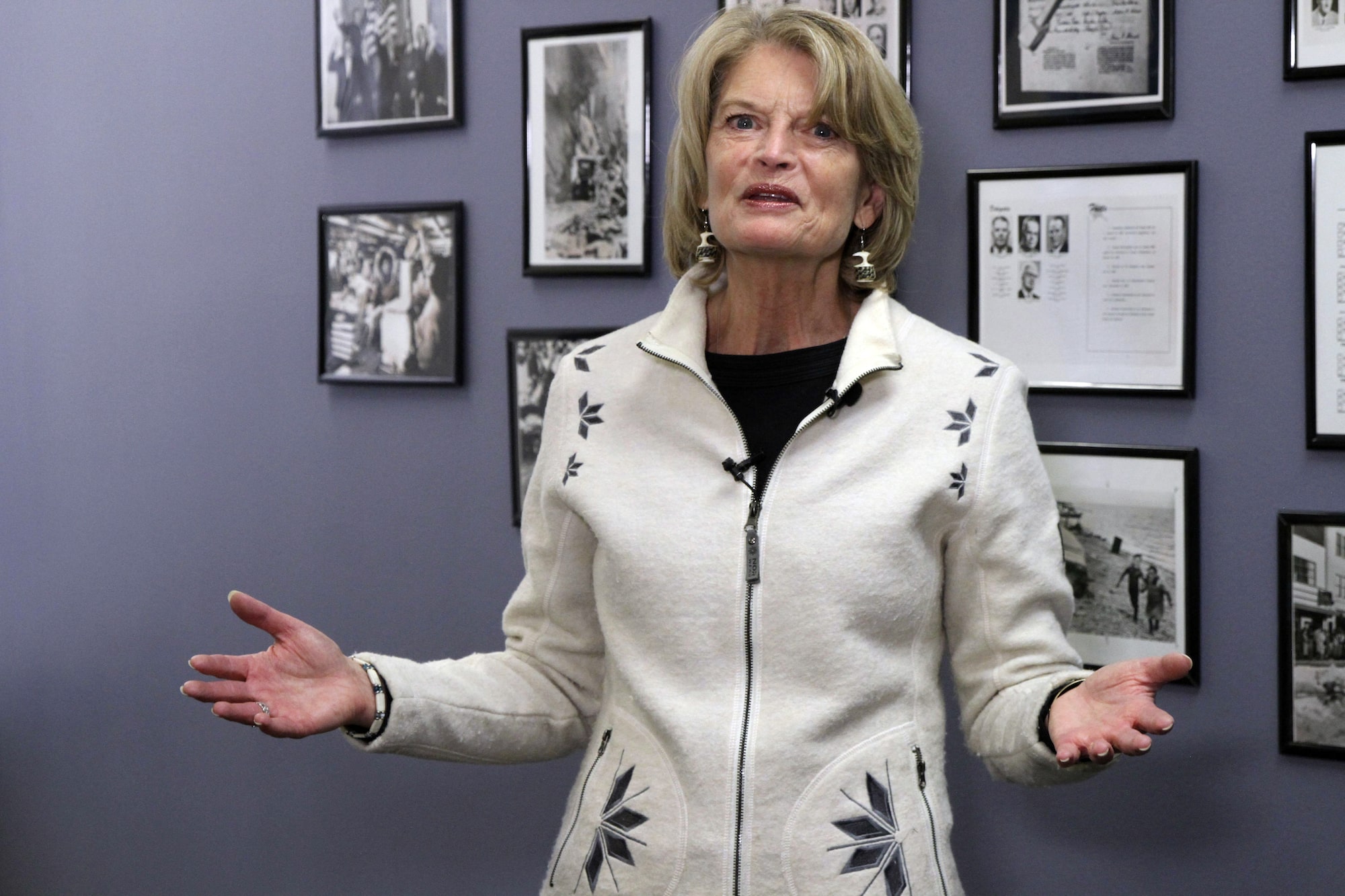 A woman in a white zip-up sweater with snowflake embroidery holds both hands out while looking at the camera. She is standing in front of a purple wall hung with black and white photos