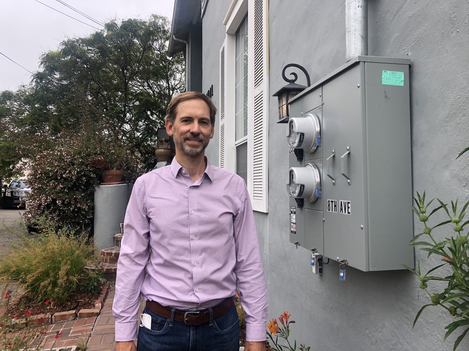 a man in a pink shirt and jeans stands outside a gray house with an electrical box on the side