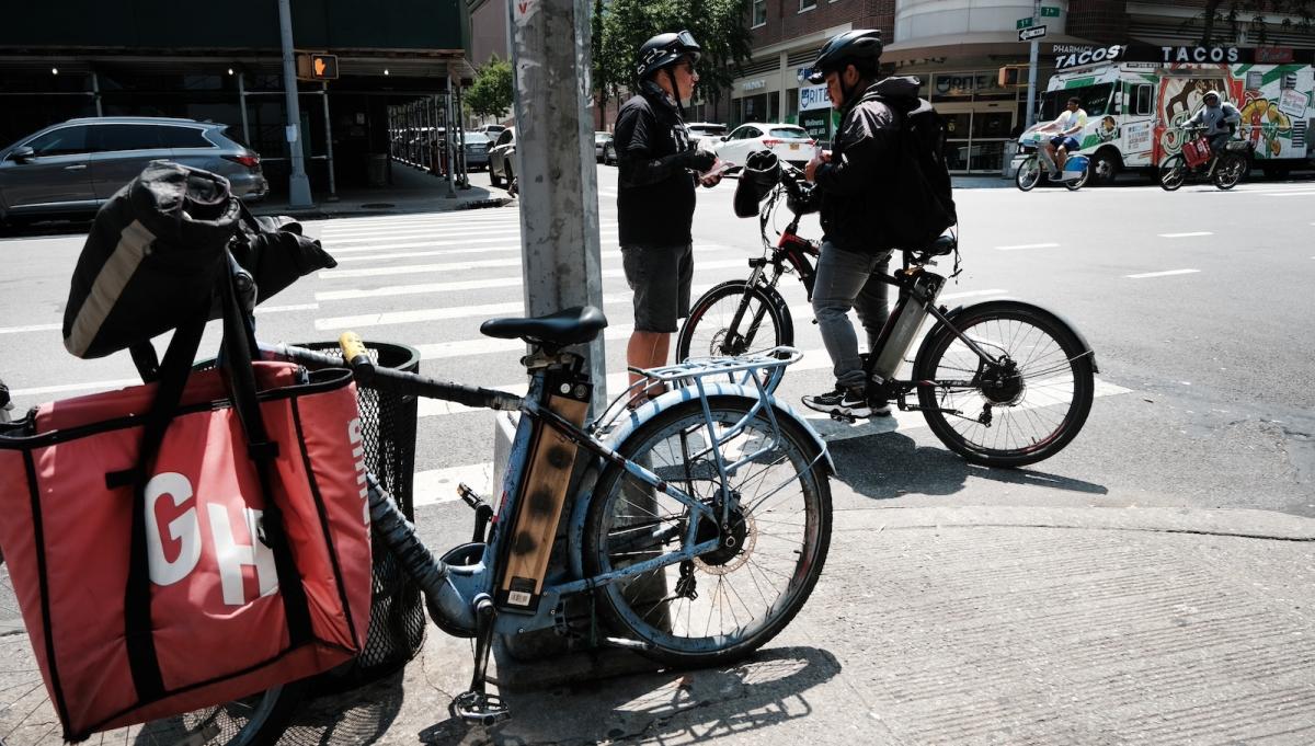 A person stands on a city street wearing a helmet and talking to another person, also helmeted, on a bike, with another bike with a red bag with the letters GH on it in the foreground