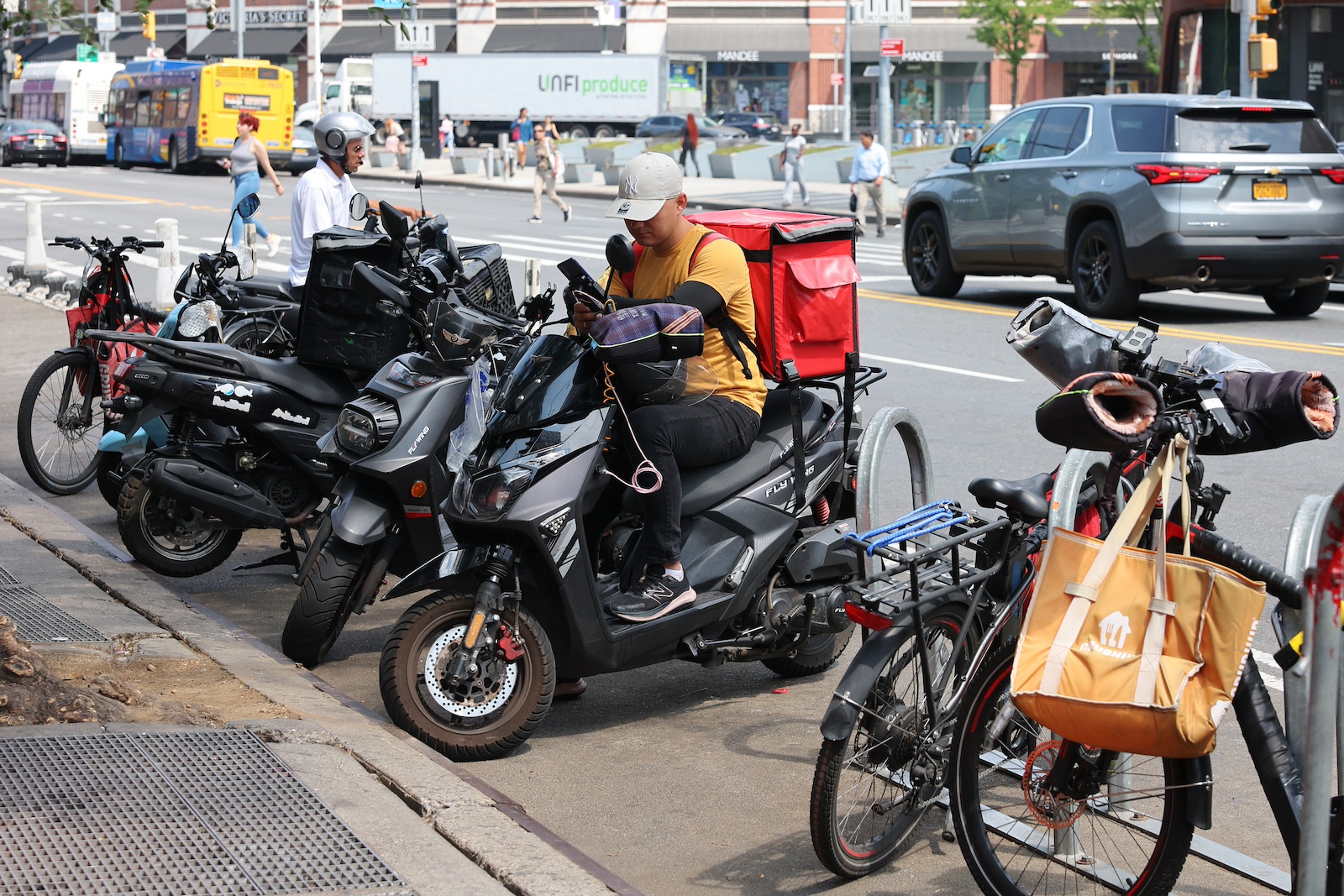 A person in an orange shirt sits on a black moped with a red cooler on the back, surrounded by other bikes and mopeds, with a city street in the background