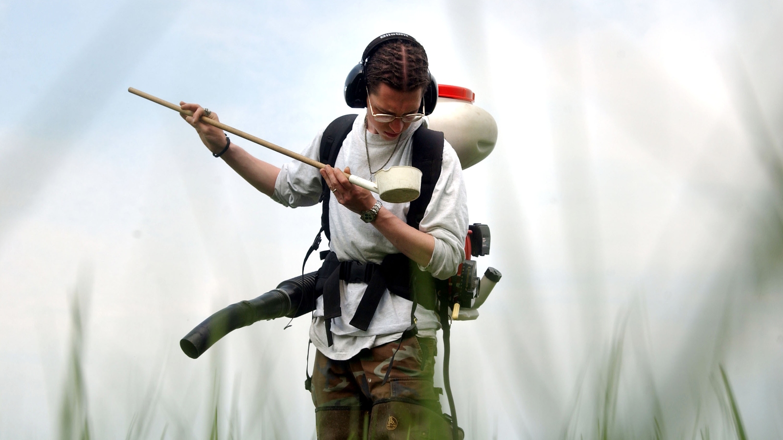 A person examines a long stick with a white cup on the end of it in a field. They are wearing a mosquito-spraying device.