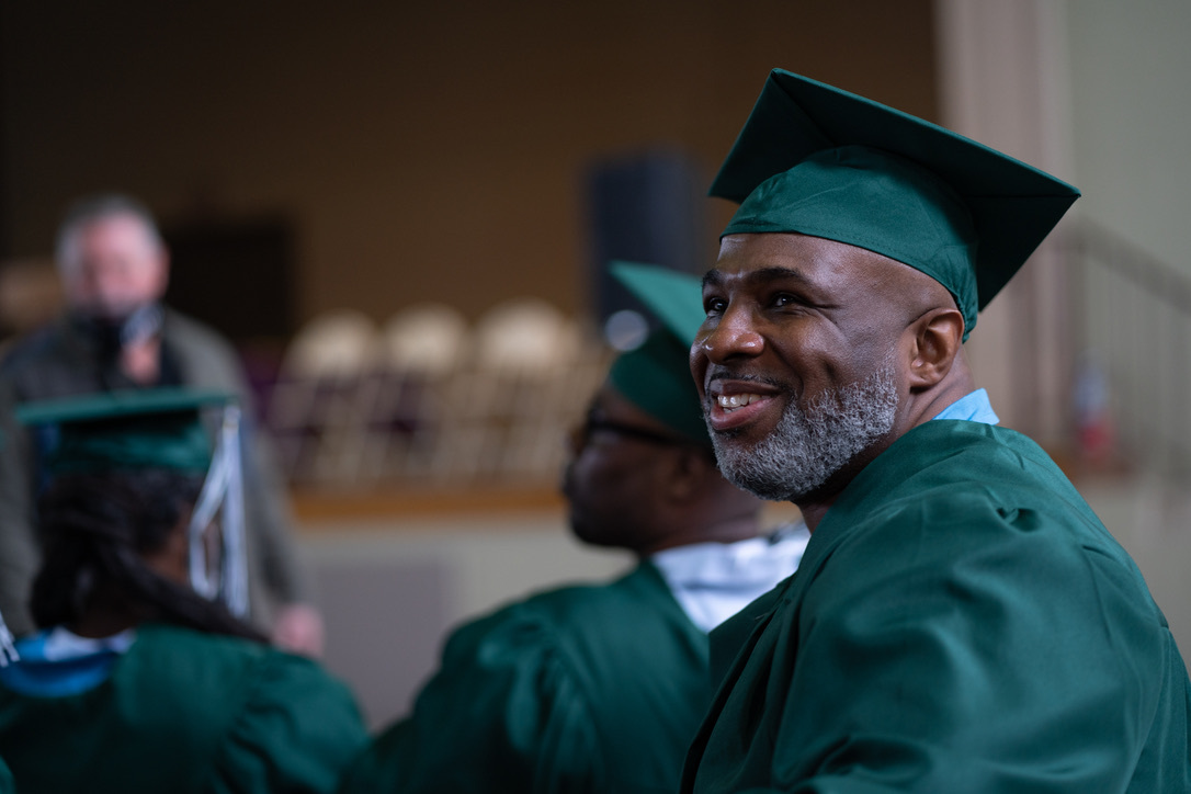 A smiling man with a gray beard wears a green cap and gown, and sits next to other graduates.