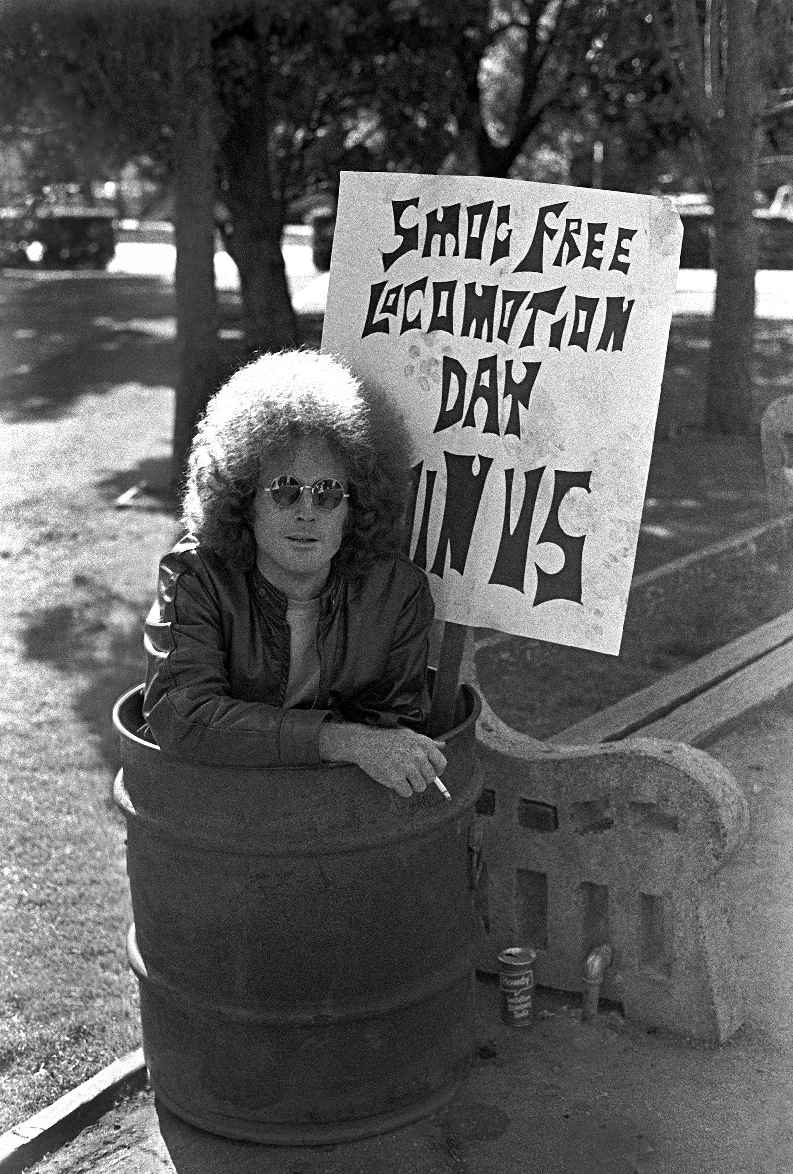 A man with long curly hair sits in a trash can holding a protest sign