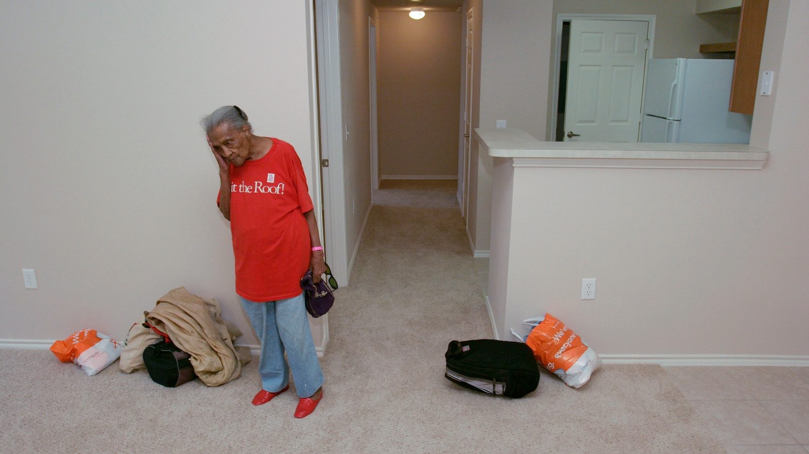 A woman stands in a hallway wearing a red shirt, her hand on her face. Next to her is a backpack and a pile of belongings.